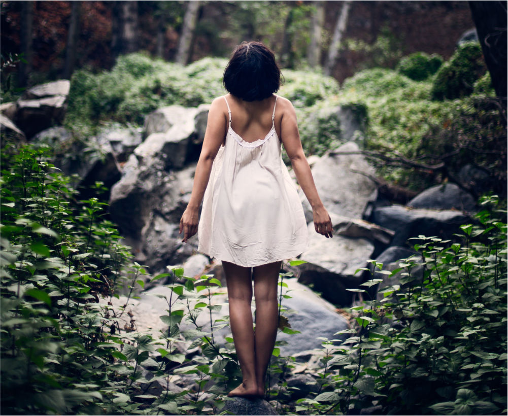 woman in white dress standing with her back towards us on a rock in a forest with slightly opened arms